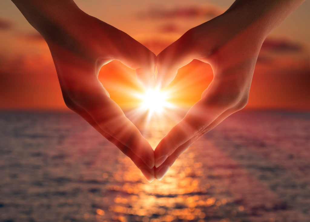 Two hands form a heart with an ocean sunset background.
