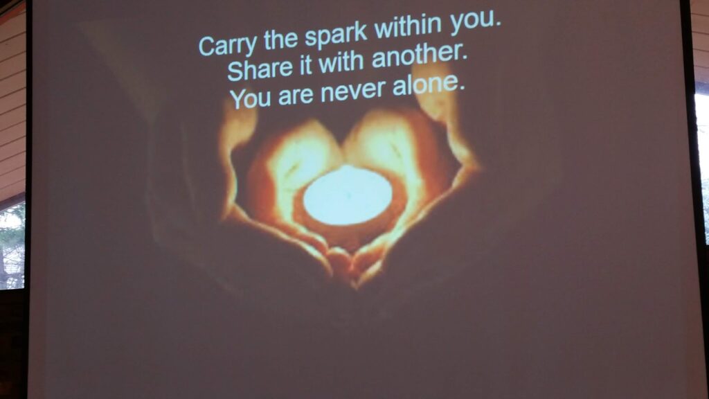 Carry the spark within you. Share it with another. You are never alone."  Over background of two sets of hands forming a heart, holding a small candle.