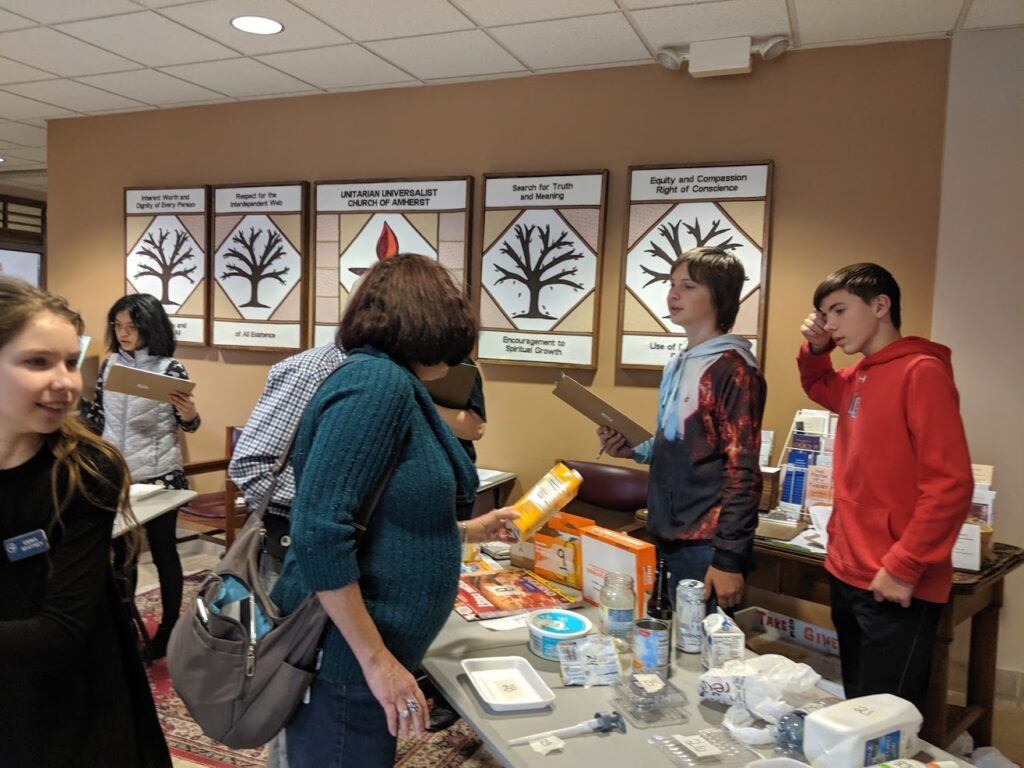Teens standing behind table filled with many types of disposable items with adults on other side picking up items to decide whether to Reduce, Recycle, Reuse.